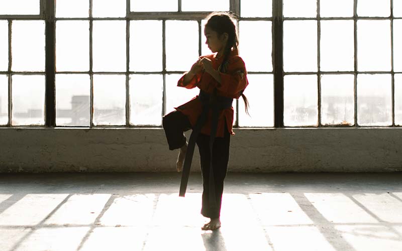 Nena vestida amb uniforme d'arts marcials davant d'una finestra molt gran amb vidres quadrats petits.